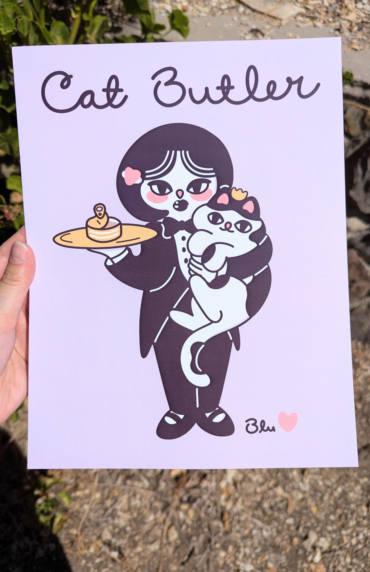 A hand holds a square art print outdoors. The print features a stylized cartoon illustration of a woman in a tuxedo holding a white cat, with the text 'Cat Butler' at the top and the signature 'Blu' with a small pink heart at the bottom.