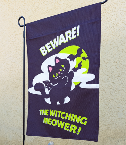 A dark purple Halloween garden flag hanging from a black metal stand against a beige stucco wall. The flag features a cartoon black cat with glowing green eyes and bat wings, standing in front of a yellow full moon and white clouds, with the text 'BEWARE! THE WITCHING MEOWER!' in white and neon green.