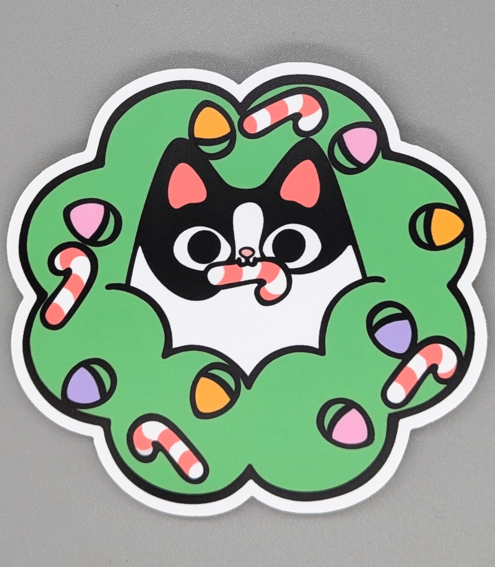 Sticker of a cat in a green wreath with candy canes on a gray background