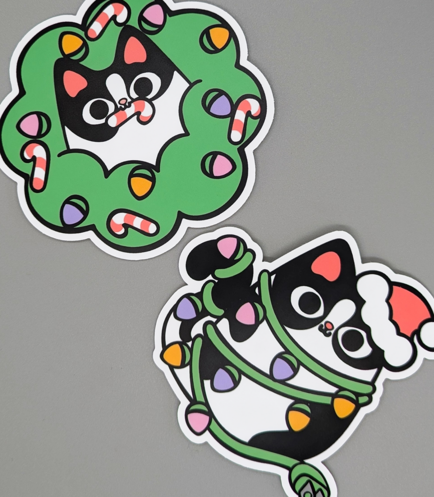 Two festive stickers of a black cat with a wreath and another with lights on a gray background.