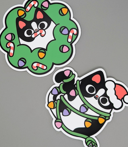 Two festive stickers of a black cat with a wreath and another with lights on a gray background.