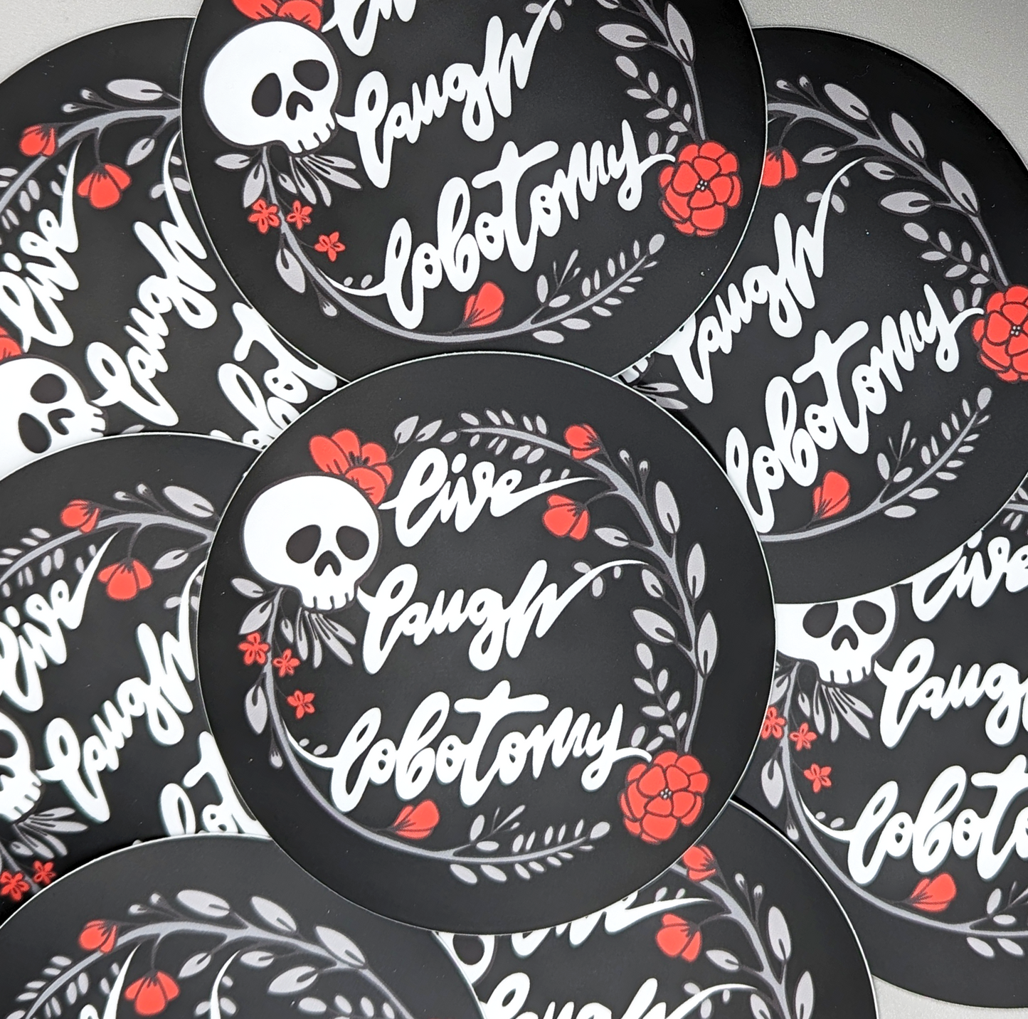 Live Laugh Lobotomy Circle Vinyl Sticker. Sticker says "Live, Laugh, Lobotomy" and features a skull and flowers design.